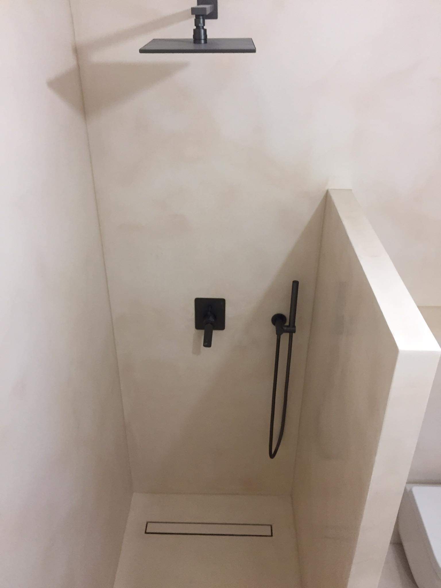 Nowoczesny prysznic typu walk-in z czarną armaturą, odpływem liniowym i ścianami wykończonymi w jasnym odcieniu beżu.