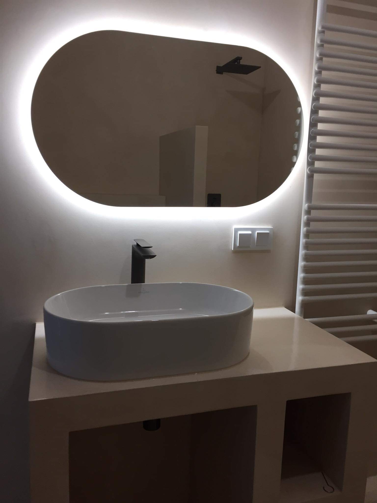 Nowoczesna łazienka z owalnym lustrem podświetlanym LED, białą umywalką nablatową z czarną baterią i grzejnikiem drabinkowym w tle.