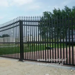 Czarna, metalowa brama wjazdowa z ostrymi zakończeniami prętów, osadzona między dwoma filarami wykończonymi imitacją kamienia łupanego, widok z zewnątrz posesji.