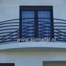 Balkon z metalową, giętą balustradą w kolorze grafitowym, umieszczony na białej elewacji budynku, z widocznym oknem za balustradą i adresem strony internetowej nałożonym na zdjęcie.