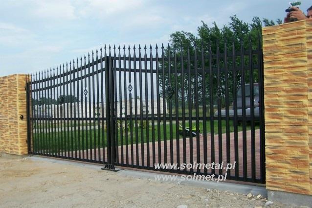 Czarna, metalowa brama wjazdowa z ostrymi zakończeniami prętów, osadzona między dwoma filarami wykończonymi imitacją kamienia łupanego, widok z zewnątrz posesji.