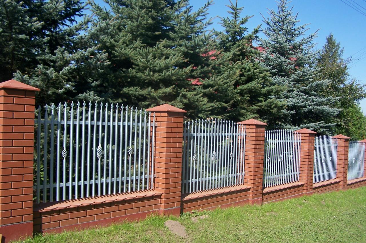 Niski murek z cegły klinkierowej zwieńczony metalowym, szarym ogrodzeniem sztachetowym z ozdobnymi elementami w kształcie liści, na tle zielonych drzew iglastych i trawnika.