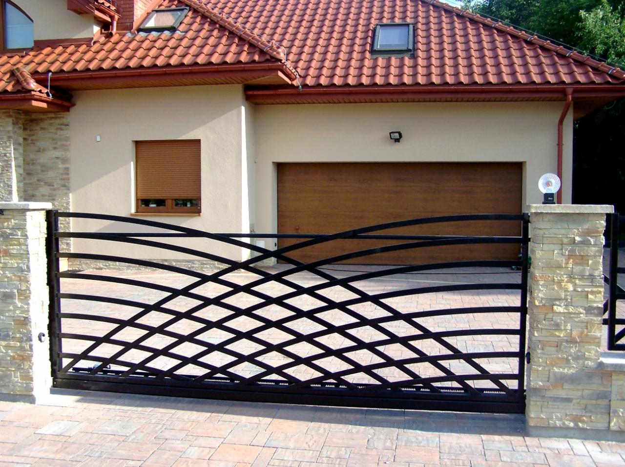 Czarna, metalowa brama wjazdowa o falującym wzorze na tle domu z beżową elewacją i brązową bramą garażową. Brama osadzona na kamiennych słupkach.