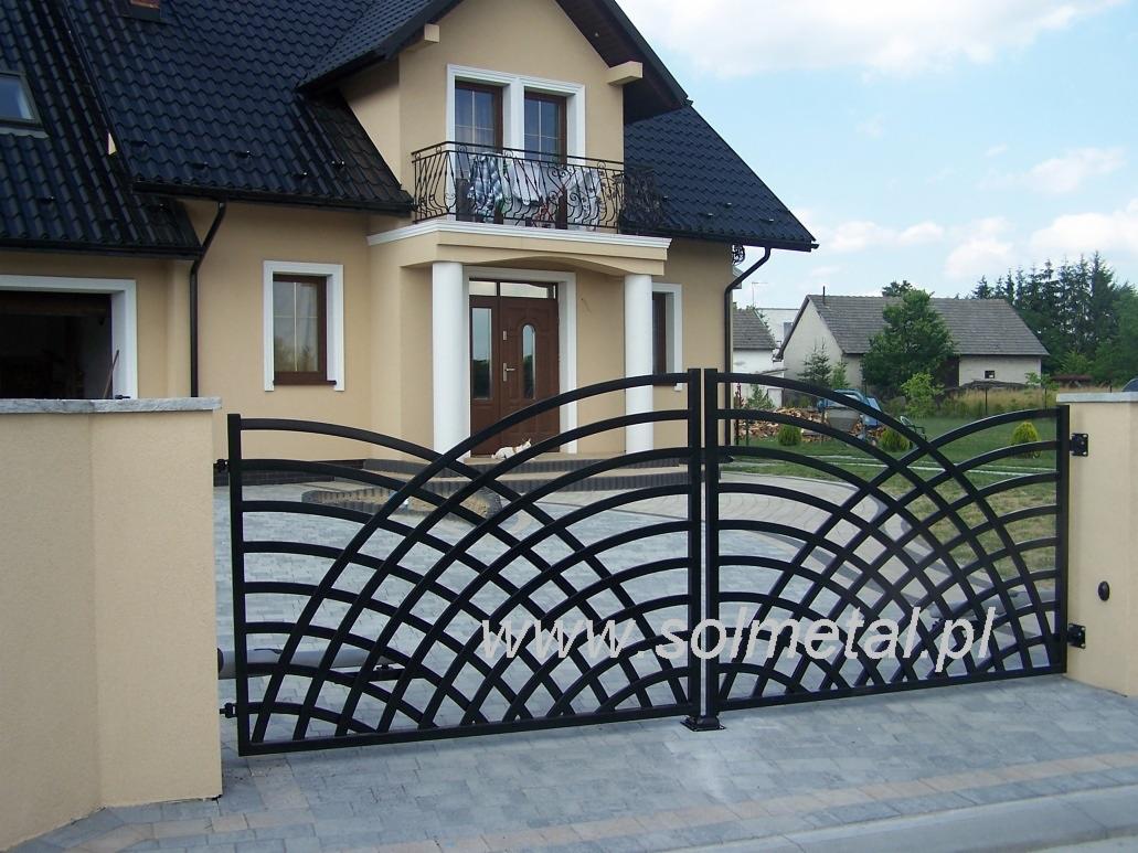 Czarna metalowa brama wjazdowa o dekoracyjnym, łukowatym wzorze kratownicy, zamontowana między beżowymi słupkami ogrodzeniowymi, na tle domu z balkonem i kostki brukowej.
