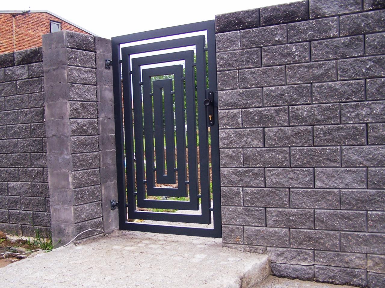 Nowoczesna furtka z metalu o geometrycznym wzorze, wkomponowana w mur z szarej cegły betonowej, z widocznym betonowym słupkiem po lewej stronie.