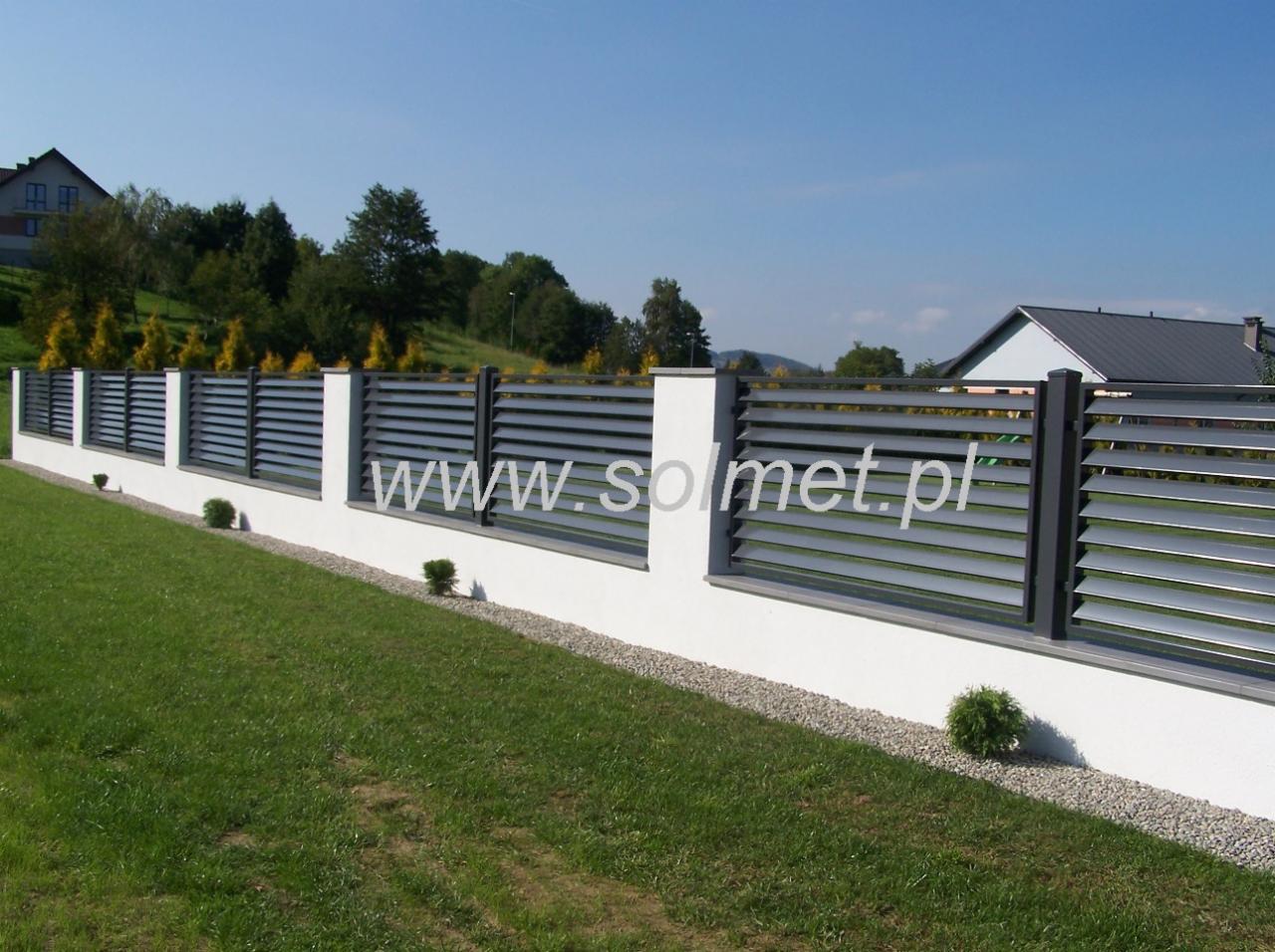 Nowoczesne ogrodzenie z poziomych, szarych paneli metalowych, osadzone na niskim, białym murku z kamiennym obramowaniem, widok na zadbany trawnik i dom w tle, z widocznym adresem strony...