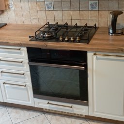 Meble Lili - Fragment zabudowy kuchennej z białymi frontami, drewnianym blatem, czarnym zlewem i płytą gazową, widoczny fragment podłogi i odkurzacza.