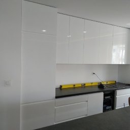 Meble Lili - Nowoczesna, biała kuchnia z błyszczącymi frontami szafek i czarnym blatem, poziomica na blacie, widoczny fragment zabudowanego piekarnika i osoba w tle.