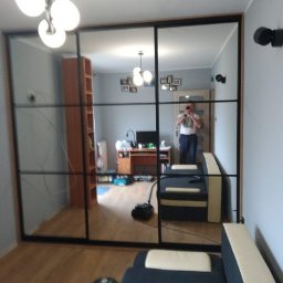 Meble Lili - Duża trzydrzwiowa szafa przesuwna z lustrzanymi frontami i czarnymi szprosami, odbijająca wnętrze pokoju w mieszkaniu w Krakowie. Widoczny mężczyzna robiący zdjęcie w lustrze.