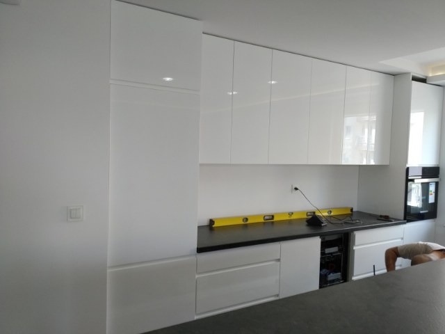 Nowoczesna, biała kuchnia z błyszczącymi frontami szafek i czarnym blatem, poziomica na blacie, widoczny fragment zabudowanego piekarnika i osoba w tle.