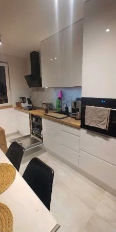 Biała, nowoczesna kuchnia z drewnianym blatem, otwartą zmywarką i czarnym okapem, widok z perspektywy jadalni.