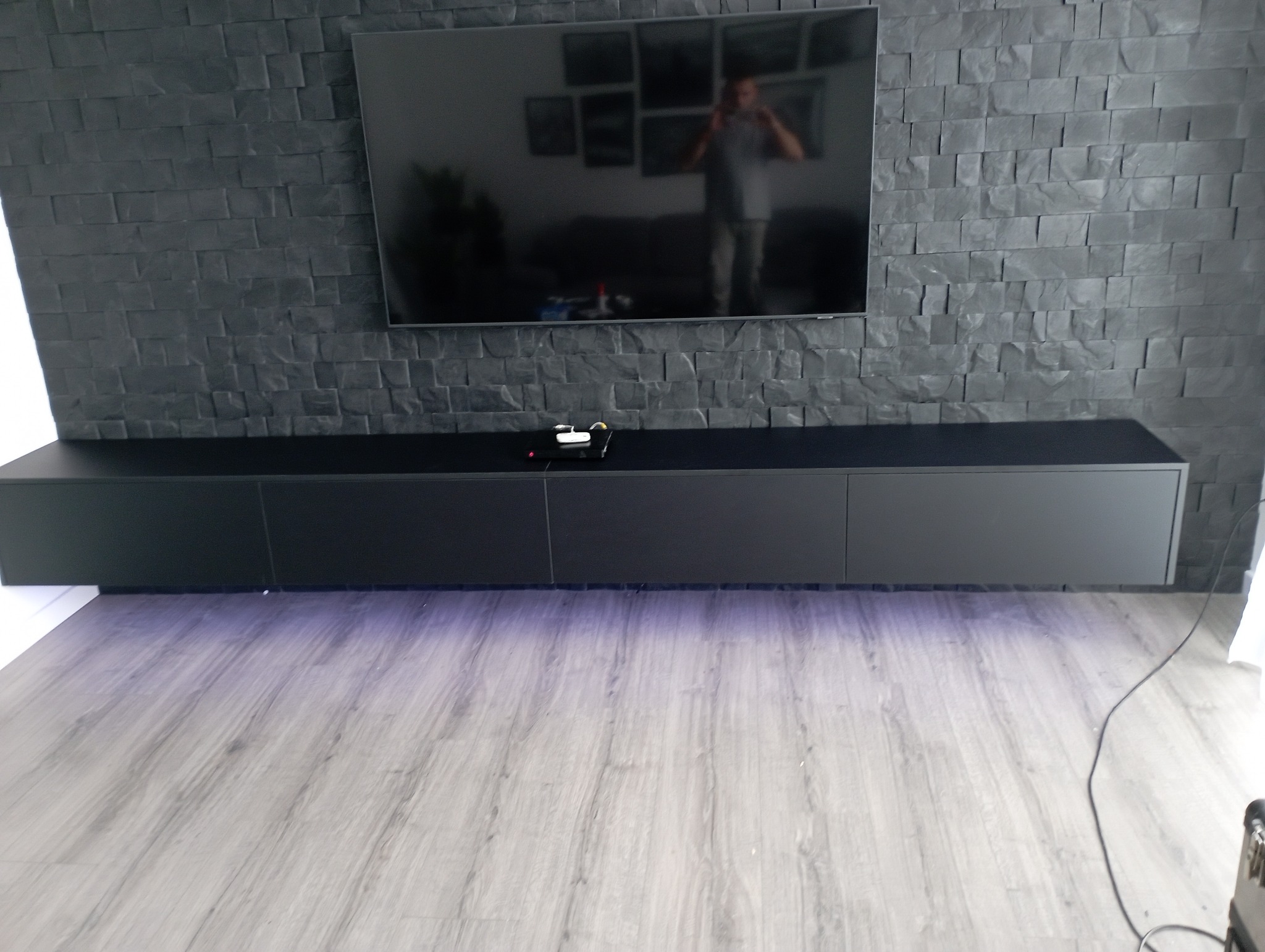 Czarna szafka RTV na wymiar, zawieszona pod telewizorem na ścianie z szarej cegły dekoracyjnej, podświetlona od dołu fioletowym światłem.