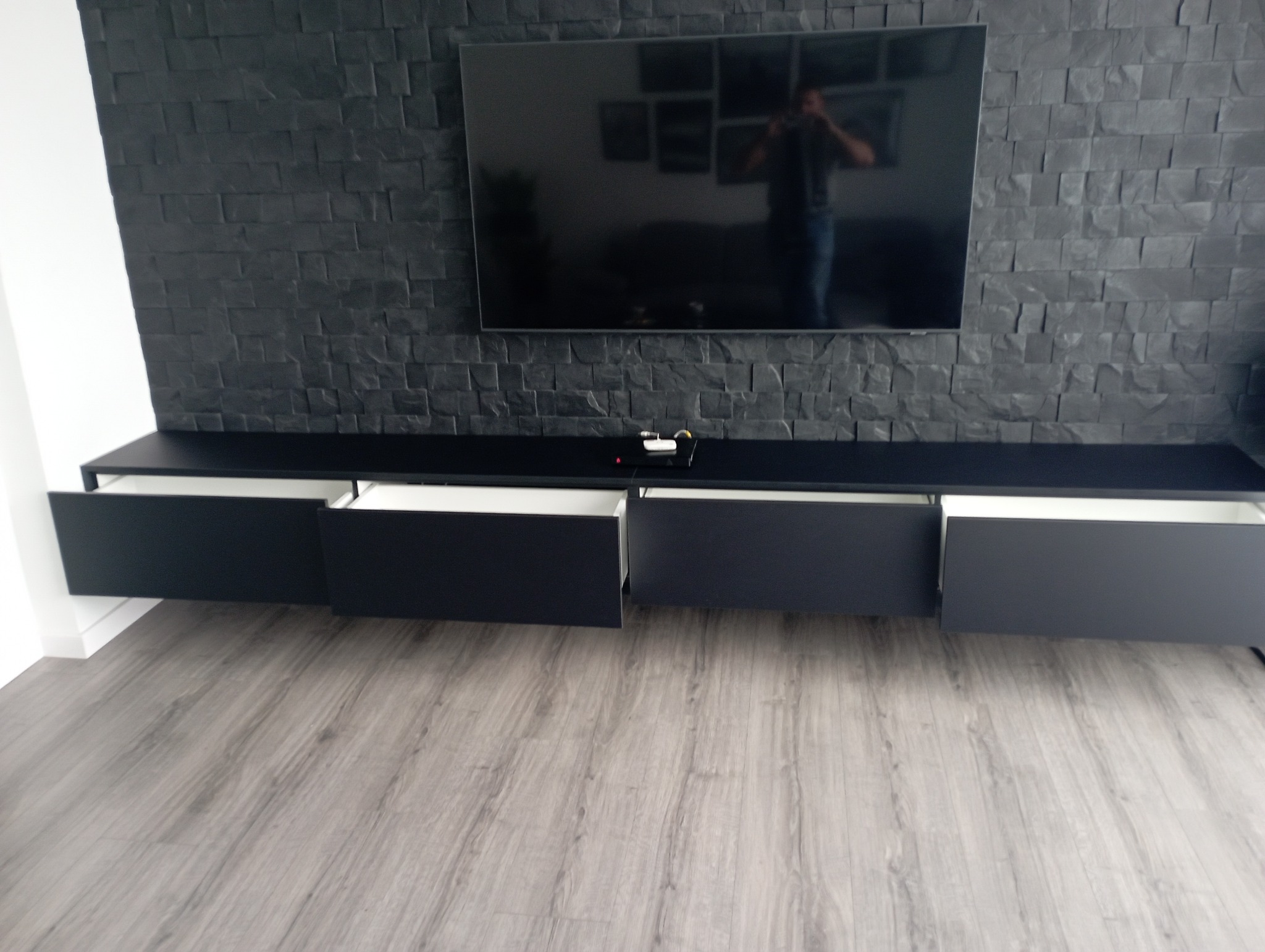 Czarna szafka RTV z otwartymi szufladami na tle ściany wyłożonej czarnym kamieniem dekoracyjnym i zawieszonego telewizora.