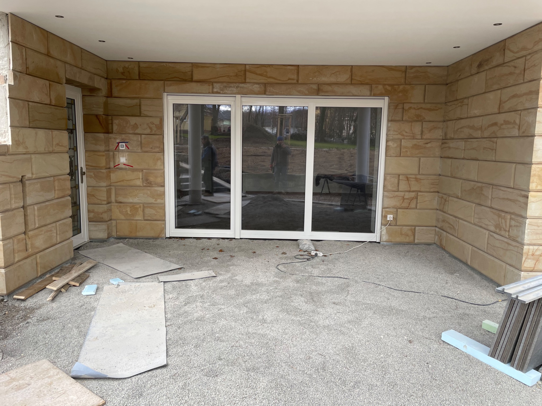 Wykończona elewacja budynku z piaskowca w trakcie prac wykończeniowych, widoczne płyty elewacyjne, okno przesuwne i materiały budowlane na podłożu.