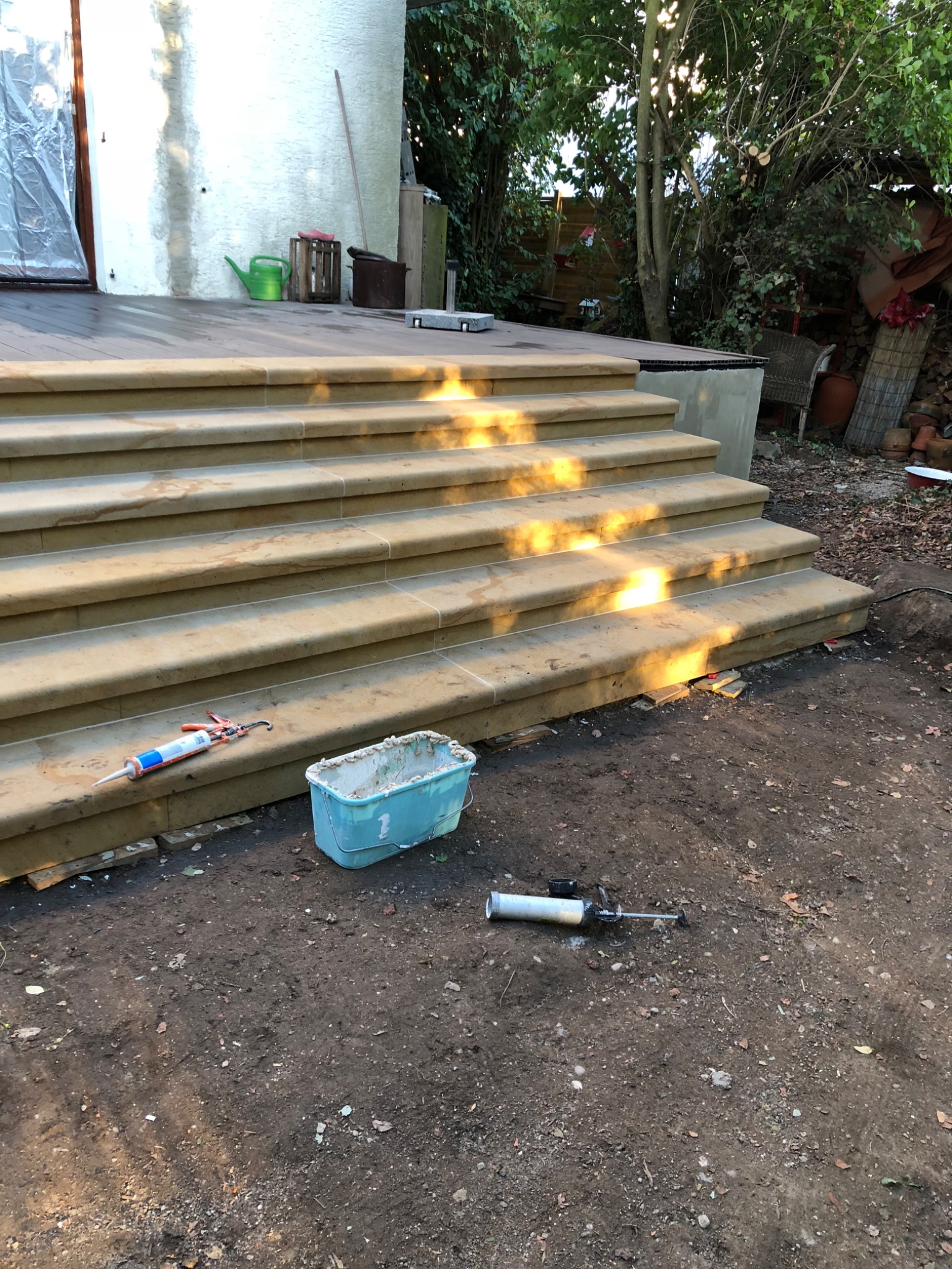 Kamienne schody zewnętrzne w trakcie budowy, widoczne narzędzia: pistolet do silikonu, wiadro z zaprawą, ziemia wokół, częściowo oświetlone słońcem.