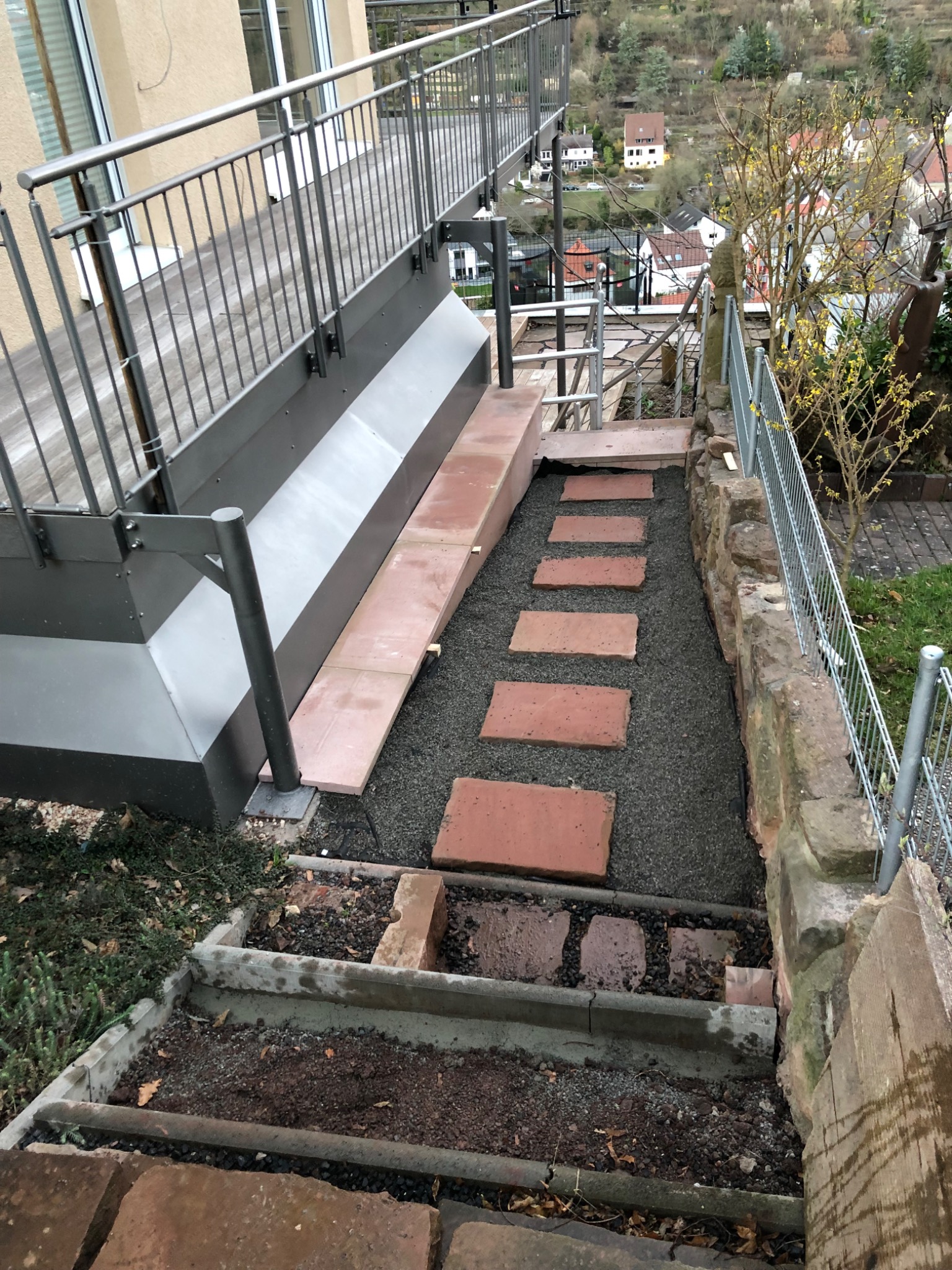 Schody zewnętrzne z metalową balustradą, wyłożone czerwonymi płytami i wysypane szarym żwirem, prowadzące w dół zbocza, widok z góry.