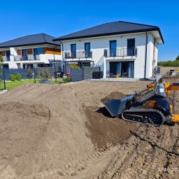 OGRODY Łukasz Gorczyca - Pomarańczowo-czarna miniładowarka Giant na placu budowy przed nowoczesnym domem z białymi ścianami i czarnym dachem, pryzma ziemi w tle, ślady opon na ubitej ziemi.