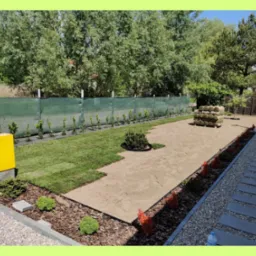 Układanie trawy z rolki w ogrodzie, widoczne fragmenty ułożonej trawy, przygotowane podłoże, rolki trawy w tle, obrzeża z roślinami i kostki chodnikowe.