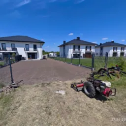 Przygotowany, wyrównany teren pod trawnik z widocznym traktorkiem glebogryzarką, w tle nowe domy jednorodzinne pod błękitnym niebem.