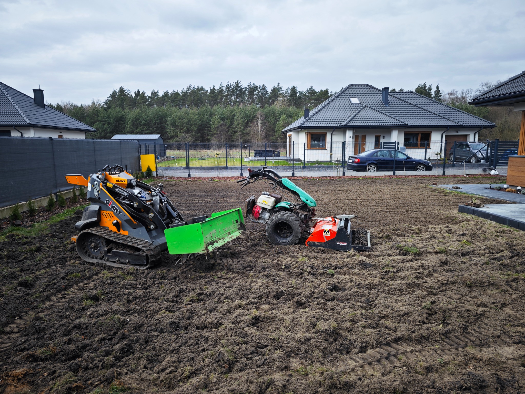 Dwa różne urządzenia do obróbki gleby na świeżo przekopanym, mokrym terenie działki z widocznymi śladami opon i budynkami mieszkalnymi w tle.