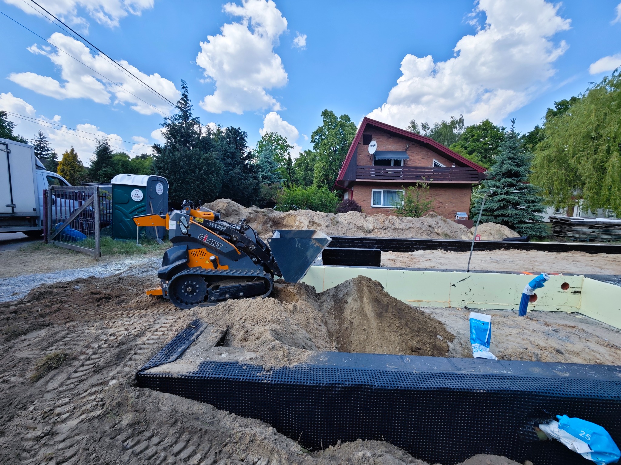Pomarańczowa miniładowarka Giant na placu budowy, wyrównująca teren pod fundamenty domu jednorodzinnego, widoczne zabezpieczenia przeciwwilgociowe, w tle przenośna toaleta i zaparkowana ciężarówka.