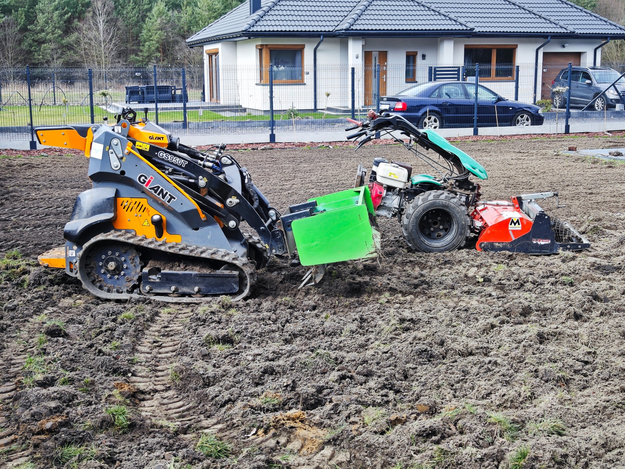 Dwie różne maszyny do przygotowywania gruntu na działce przed domem jednorodzinnym. Jedna to minitraktor gąsienicowy Giant GS950T w kolorze pomarańczowo-szarym, a druga to glebogryzarka Marathon...