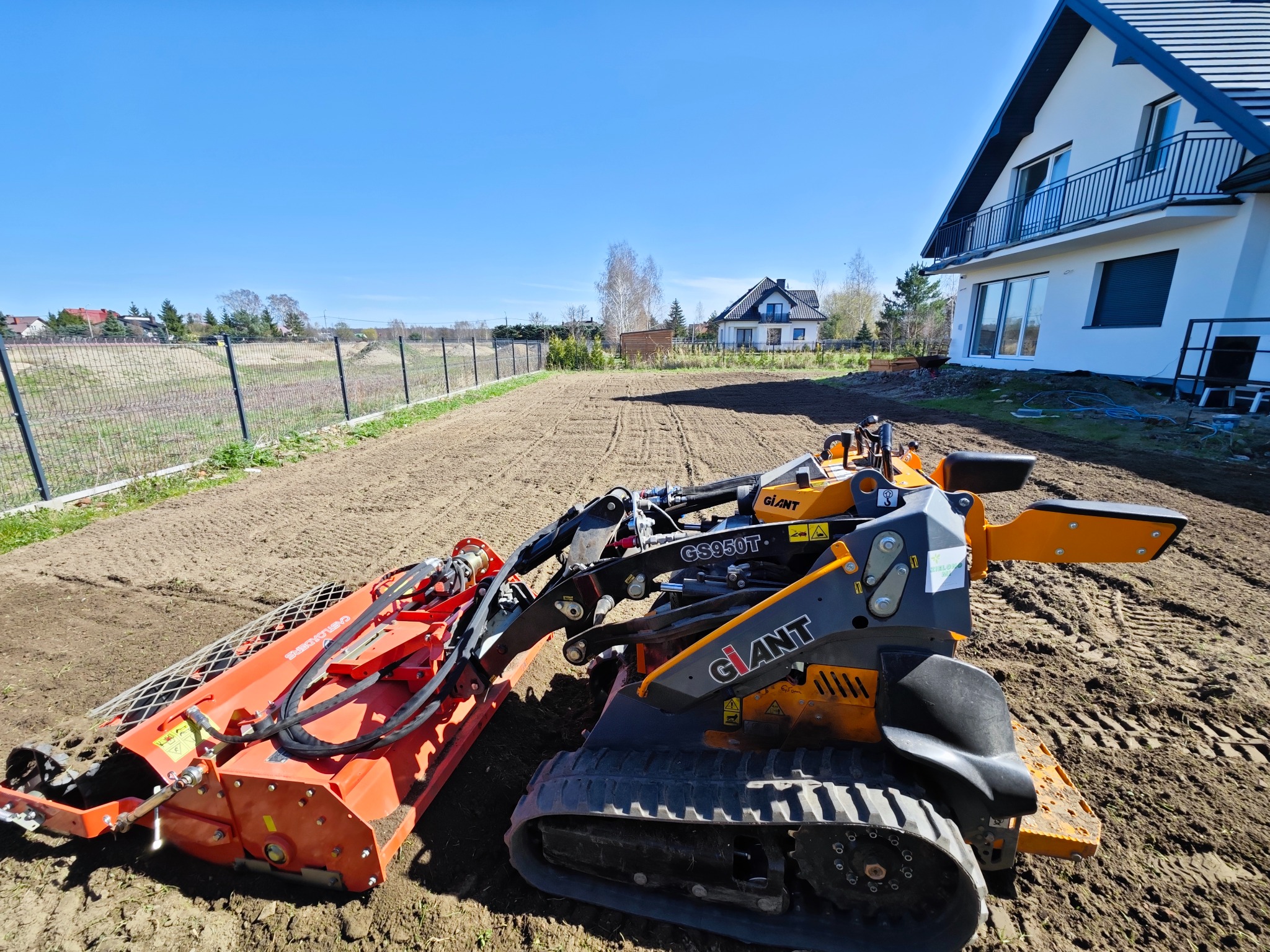 Pomarańczowo-szara frezarka glebowa marki Giant GS950T przygotowuje ziemię pod trawnik na działce z widokiem na dom jednorodzinny i ogrodzenie.