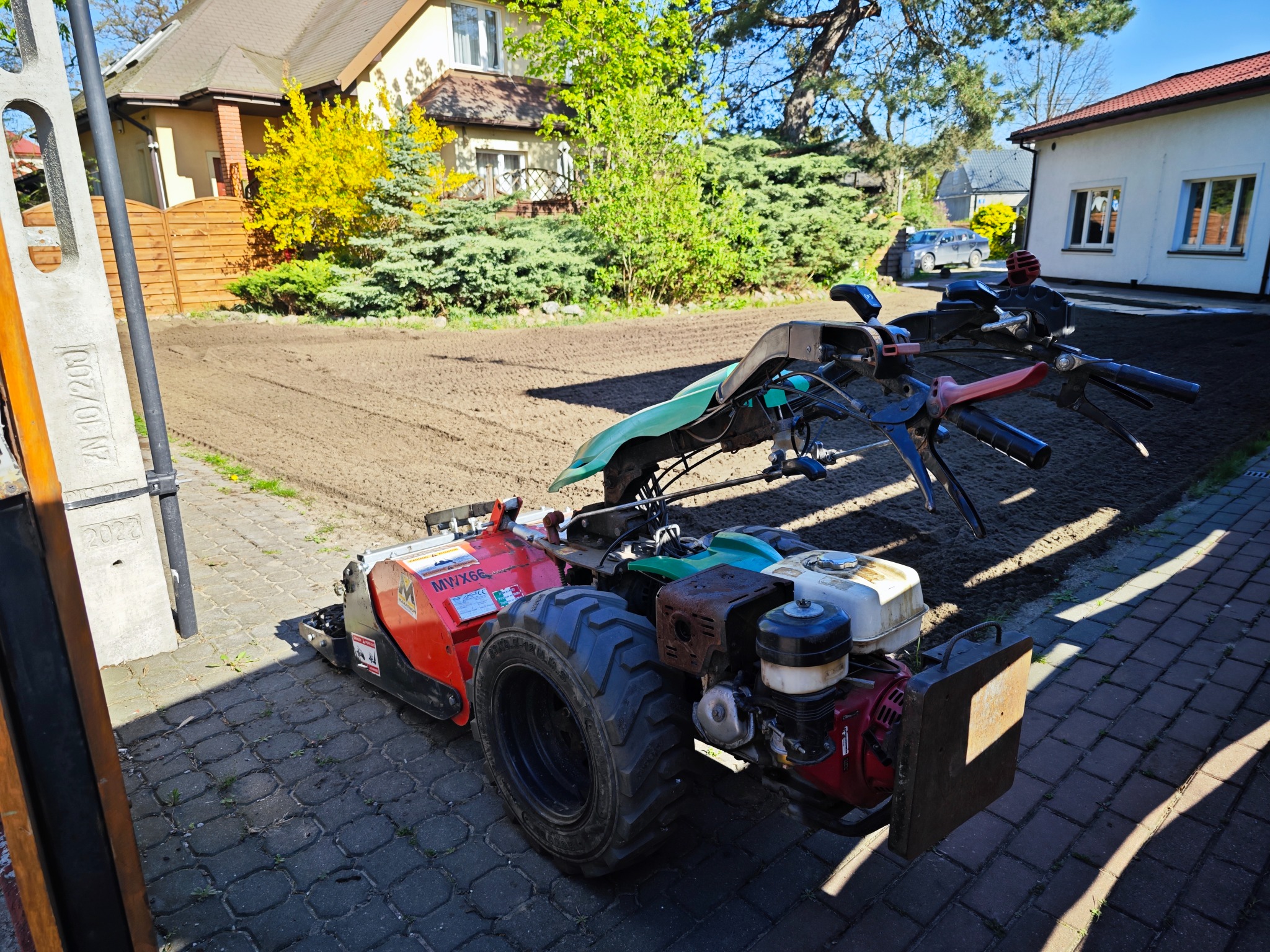 Glebogryzarka spalinowa na kostce brukowej, przygotowany teren pod trawnik w tle, domy jednorodzinne w oddali.
