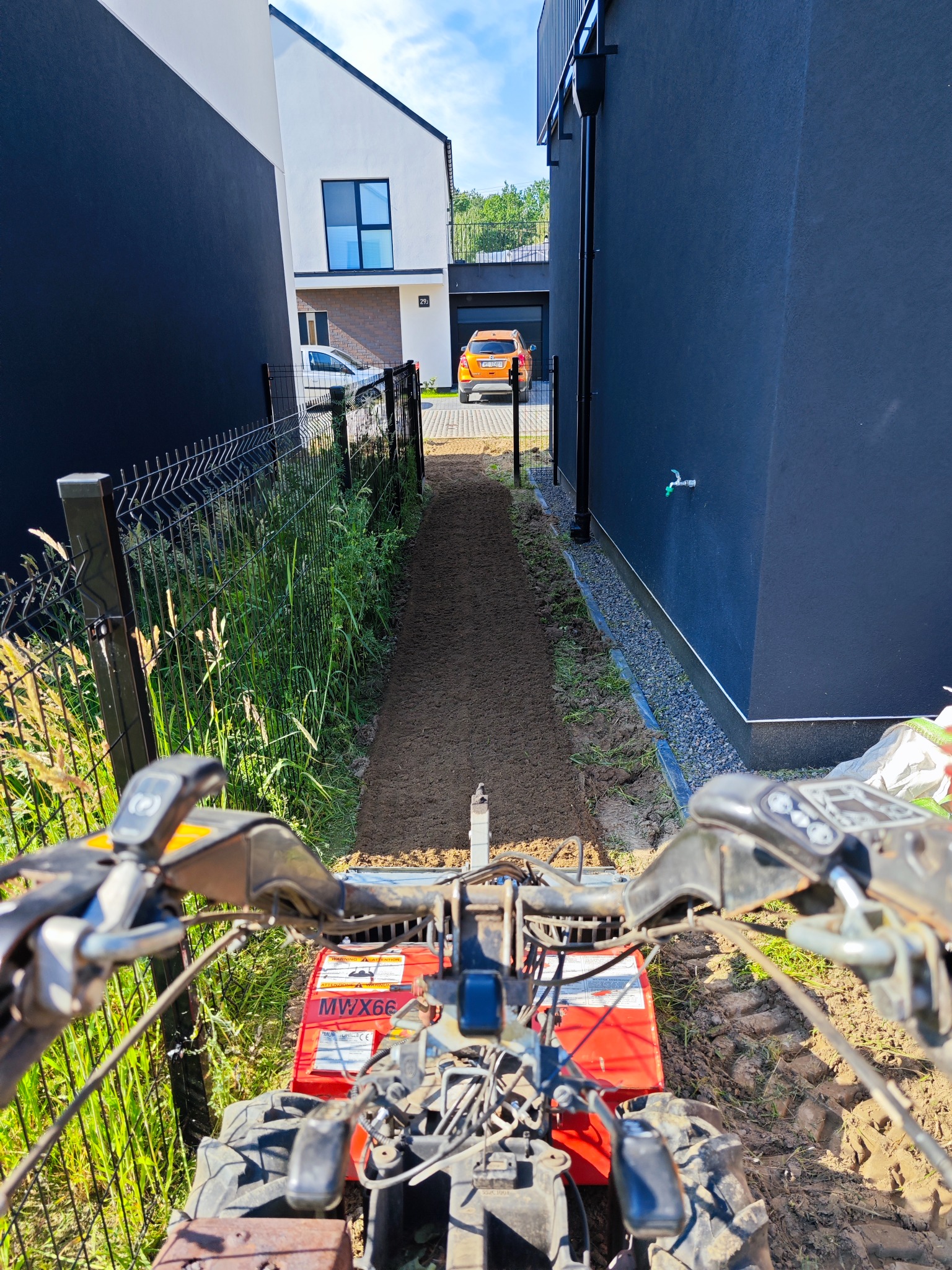 Widok z perspektywy operatora na glebogryzarkę MWMX66, przygotowującą wąski pas ziemi między nowoczesnymi domami z ciemną elewacją i metalowym ogrodzeniem.