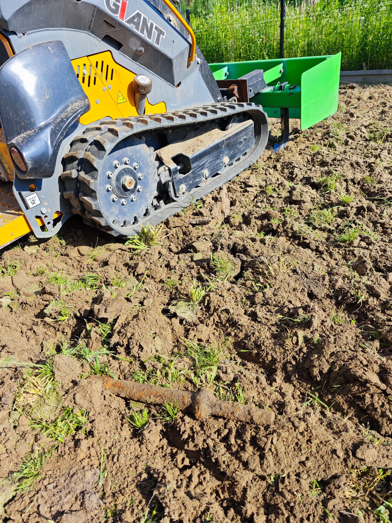 Mała gąsienicowa maszyna budowlana marki Giant z zielonym osprzętem spulchnia glebę na działce, na ziemi leży zardzewiały metalowy element.