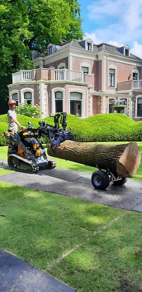 Operator w kasku transportuje dużą kłodę drewna przy użyciu miniładowarki z chwytakiem i przyczepki, na tle zadbanego ogrodu i eleganckiego domu.