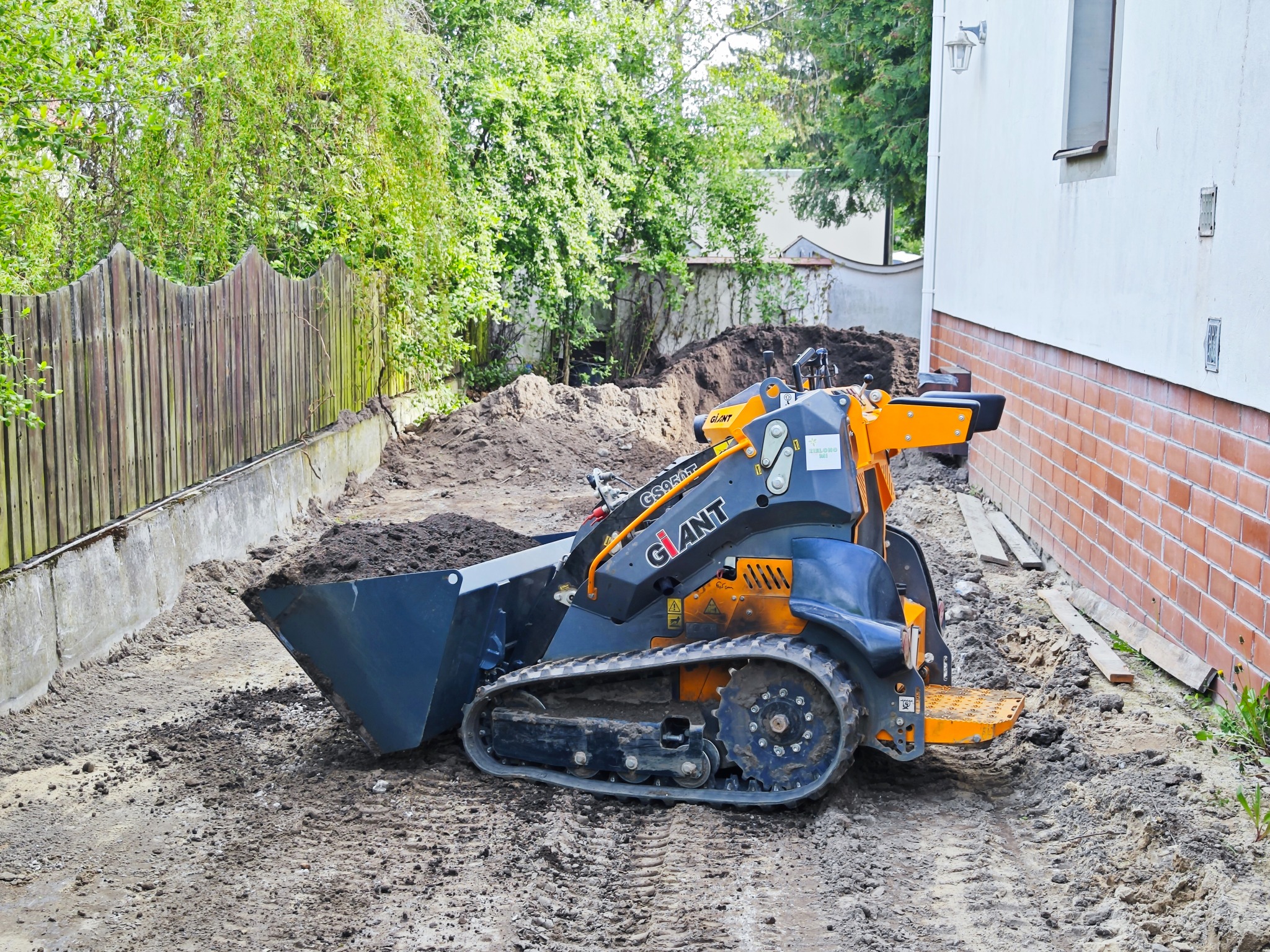 Mała, pomarańczowo-szara ładowarka gąsienicowa marki Giant stoi na ubitej ziemi obok drewnianego płotu i ściany budynku z ceglaną podmurówką, przygotowując teren pod budowę.