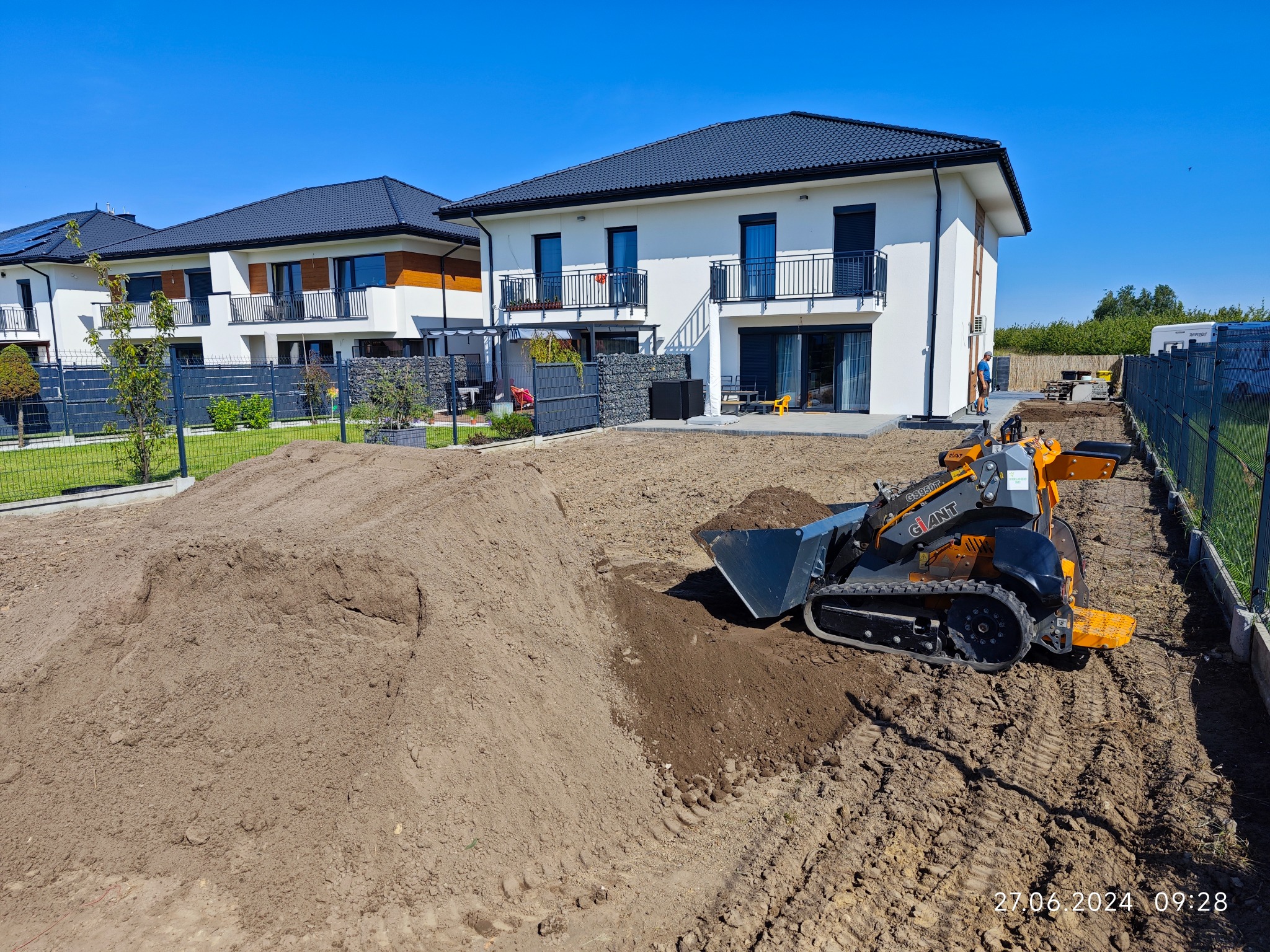 Pomarańczowo-czarna miniładowarka Giant na placu budowy przed nowoczesnym domem z białymi ścianami i czarnym dachem, pryzma ziemi w tle, ślady opon na ubitej ziemi.