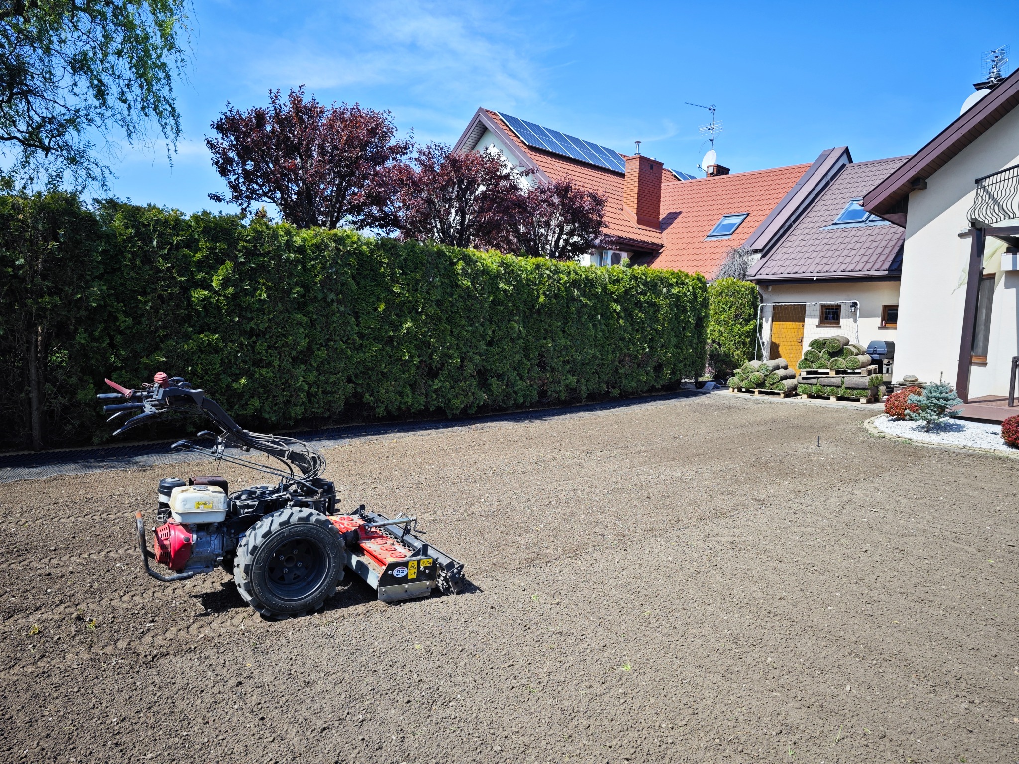 Gleba przygotowana pod trawnik z rolki, widoczna glebogryzarka spalinowa, w tle dom z panelami słonecznymi i zwinięta trawa na paletach.