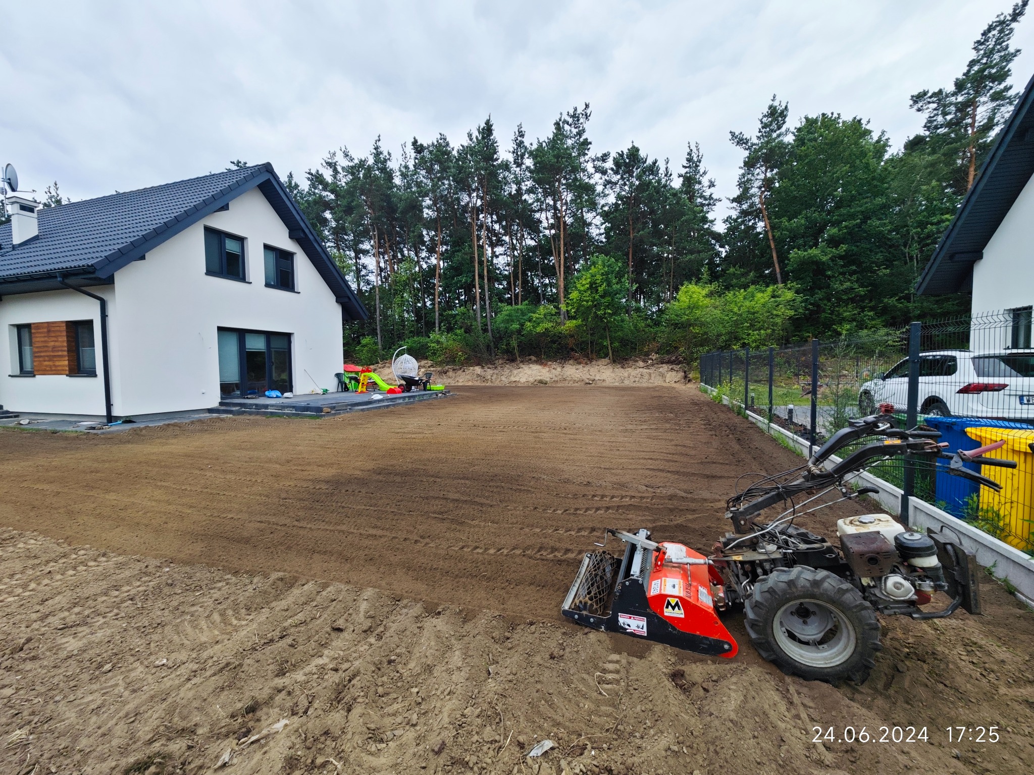 Przygotowany teren pod trawnik z widocznymi śladami po glebogryzarce, obok nowoczesny dom z ciemnym dachem i ogrodzenie panelowe.