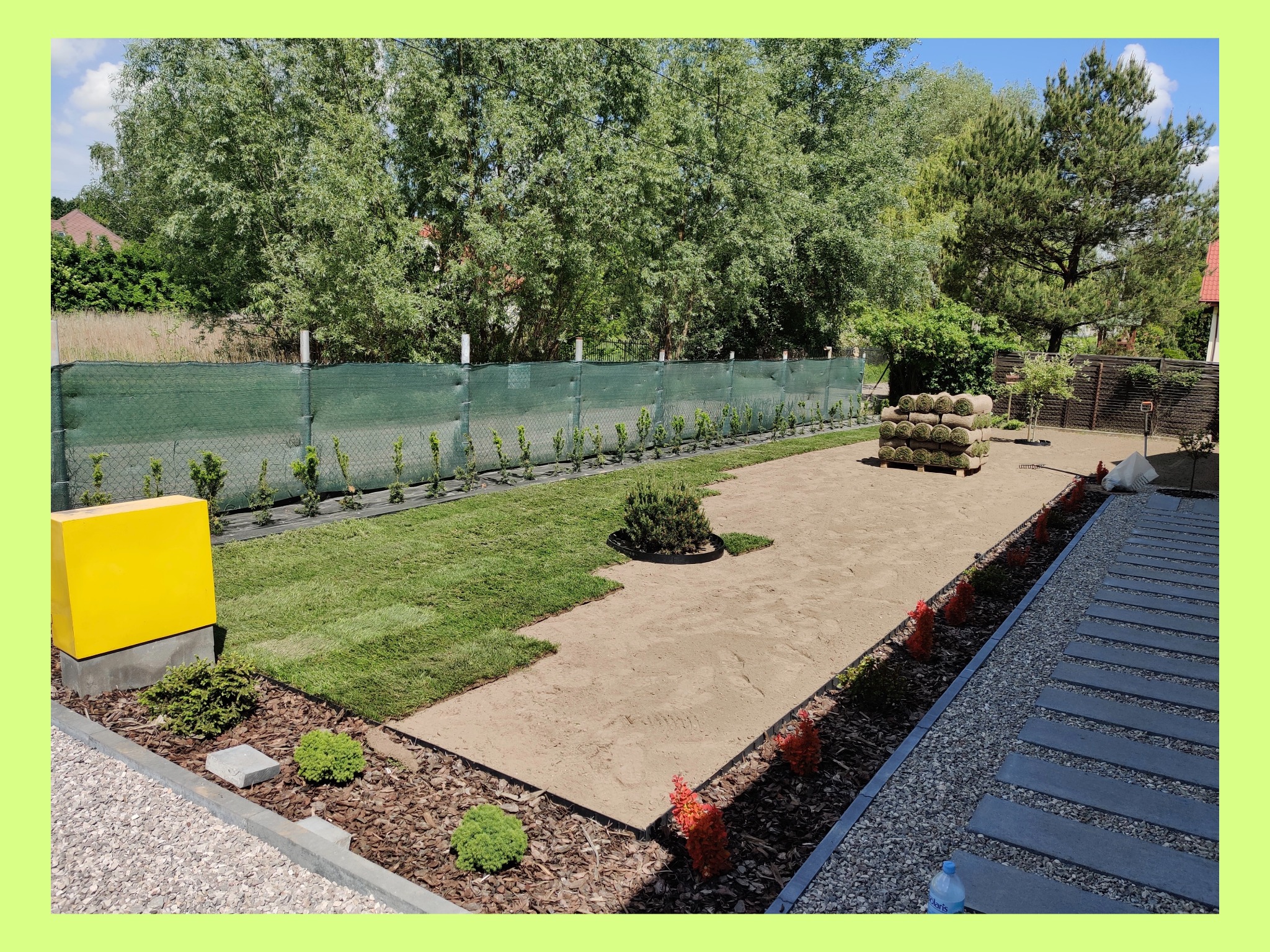 Układanie trawy z rolki w ogrodzie, widoczne fragmenty ułożonej trawy, przygotowane podłoże, rolki trawy w tle, obrzeża z roślinami i kostki chodnikowe.