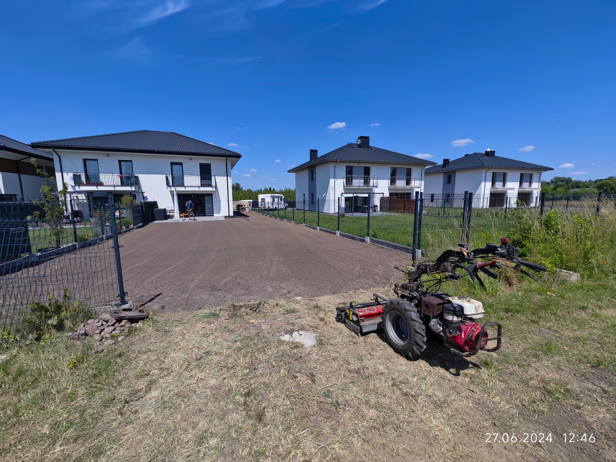 Przygotowany, wyrównany teren pod trawnik z widocznym traktorkiem glebogryzarką, w tle nowe domy jednorodzinne pod błękitnym niebem.