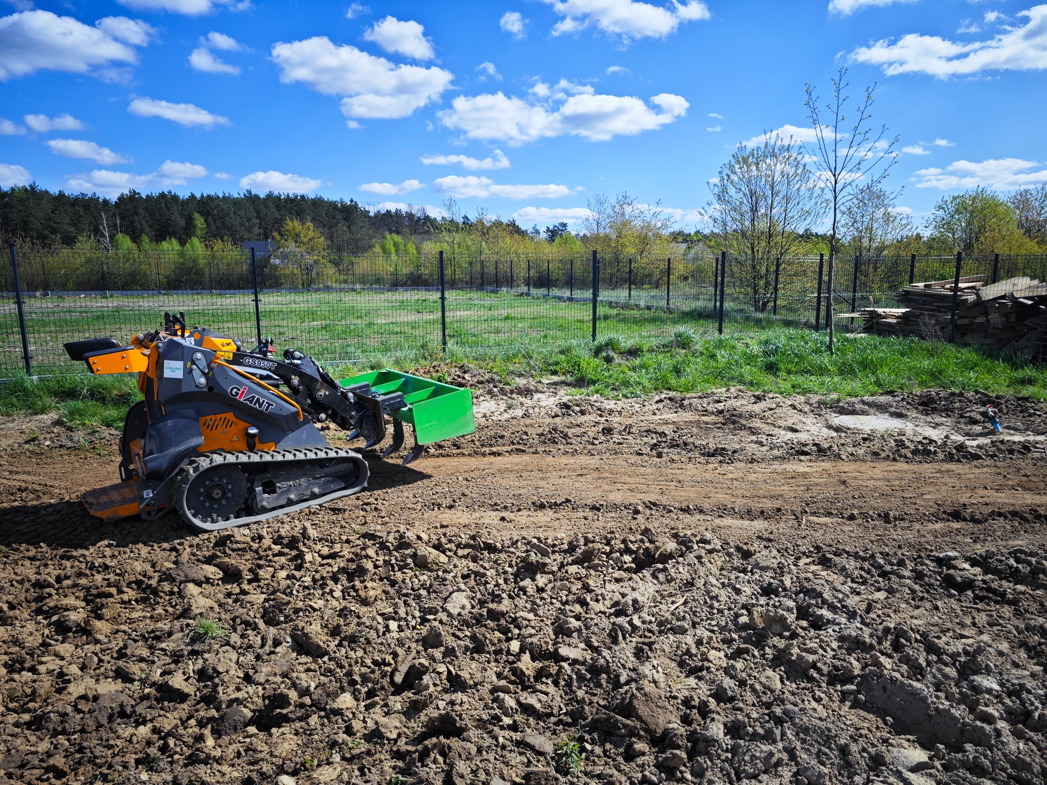 Pomarańczowo-czarna minikoparka gąsienicowa Giant z zieloną łyżką, pracująca na nierównym, brązowym terenie z widocznymi śladami opon, w tle ogrodzenie siatkowe, drzewa i błękitne niebo z chmurami.