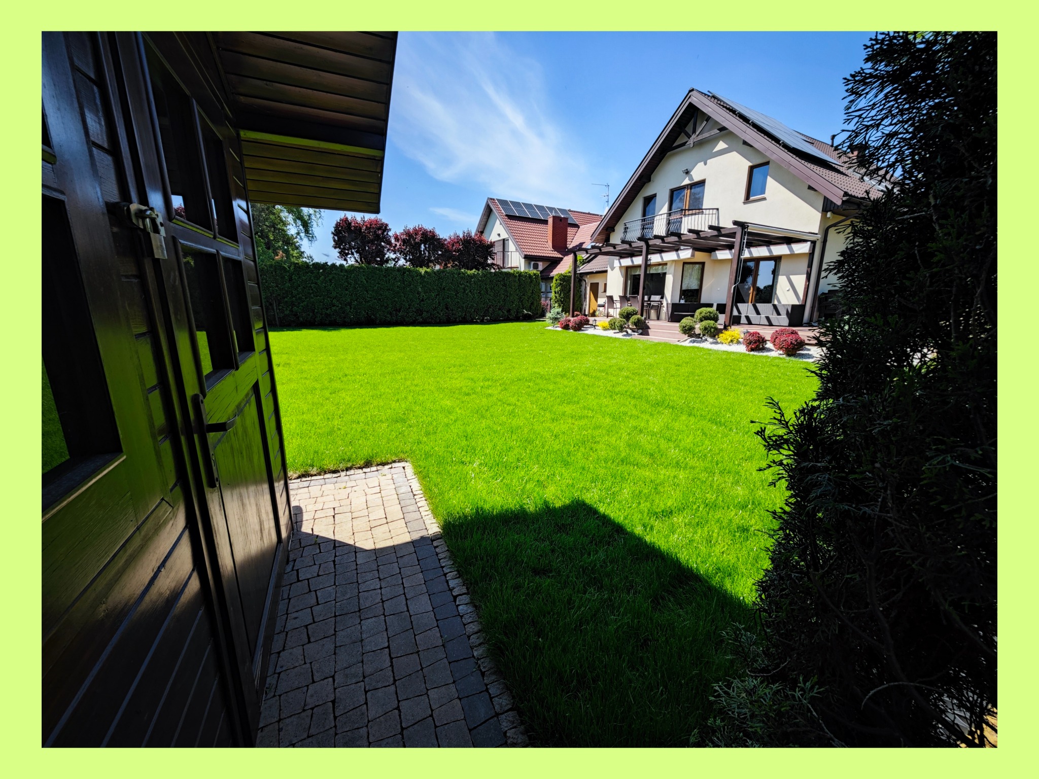 Zadbany trawnik przed domem z pergolą i tarasem, widok z perspektywy otwartej drewnianej szopy na tle błękitnego nieba.