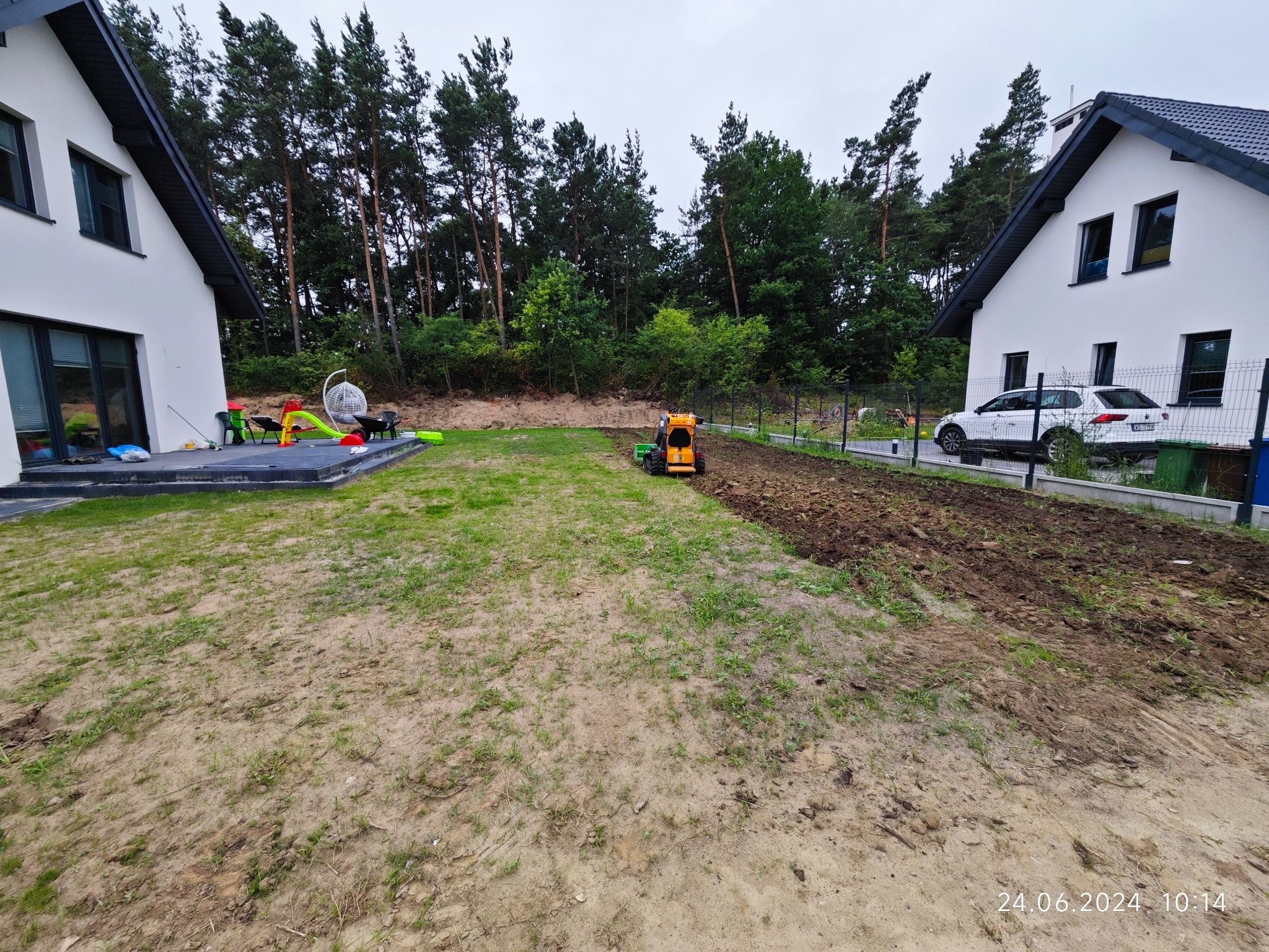 Żółta minikoparka na trawniku pomiędzy dwoma nowoczesnymi domami podczas niwelacji terenu pod przyszły ogród, widoczne świeżo przekopane pasy ziemi.