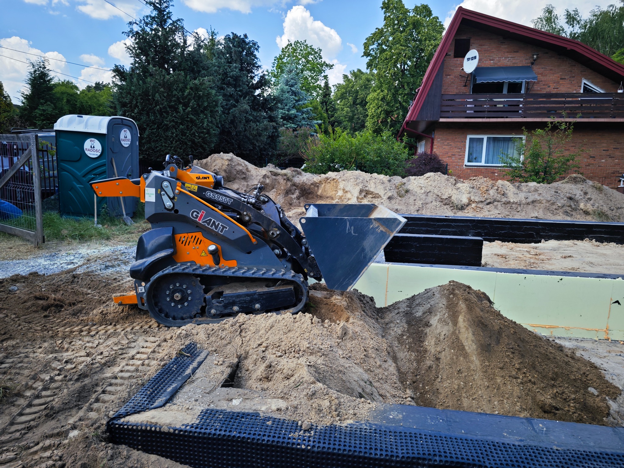 Pomarańczowa mini ładowarka gąsienicowa marki Giant, model CS950T, podczas prac ziemnych na nierównym terenie z widocznym fundamentem budynku w tle, obok przenośna toaleta