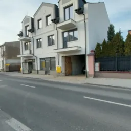 Nowoczesny, trzypiętrowy budynek z balkonami i garażem w bryle, wykończony w odcieniach bieli i szarości, widoczny z perspektywy ulicy.