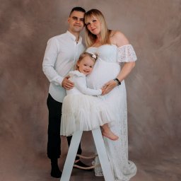 Studio_fotoradar - Uśmiechnięta rodzina: tata obejmuje mamę w ciąży, a córka siedzi na białej drabince, wszyscy ubrani na biało na tle abstrakcyjnego tła.
