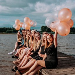 Studio_fotoradar - Grupa młodych kobiet siedzi na drewnianym pomoście nad wodą, trzymając różowe i przezroczyste balony, ubrane w sukienki i wianki z kwiatów.