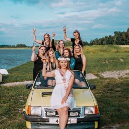 Studio_fotoradar - Grupa kobiet świętuje wieczór panieński, stojąc i siedząc na żółtym samochodzie na tle jeziora i pochmurnego nieba, trzymając kieliszki z szampanem.