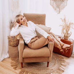 Studio_fotoradar - Uśmiechnięta blondynka w białej koszuli i beżowych spodniach, siedząca w beżowym fotelu na tle jutowej okrągłej wycieraczki, vintage walizki i dekoracji boho.