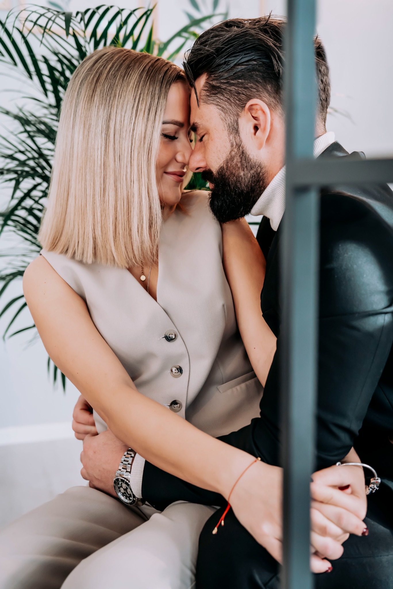 Zakochana para w Mielcu, stykająca się czołami, ona w beżowej kamizelce, on w czarnym garniturze i golfie, delikatne uśmiechy, w tle zielona roślina.