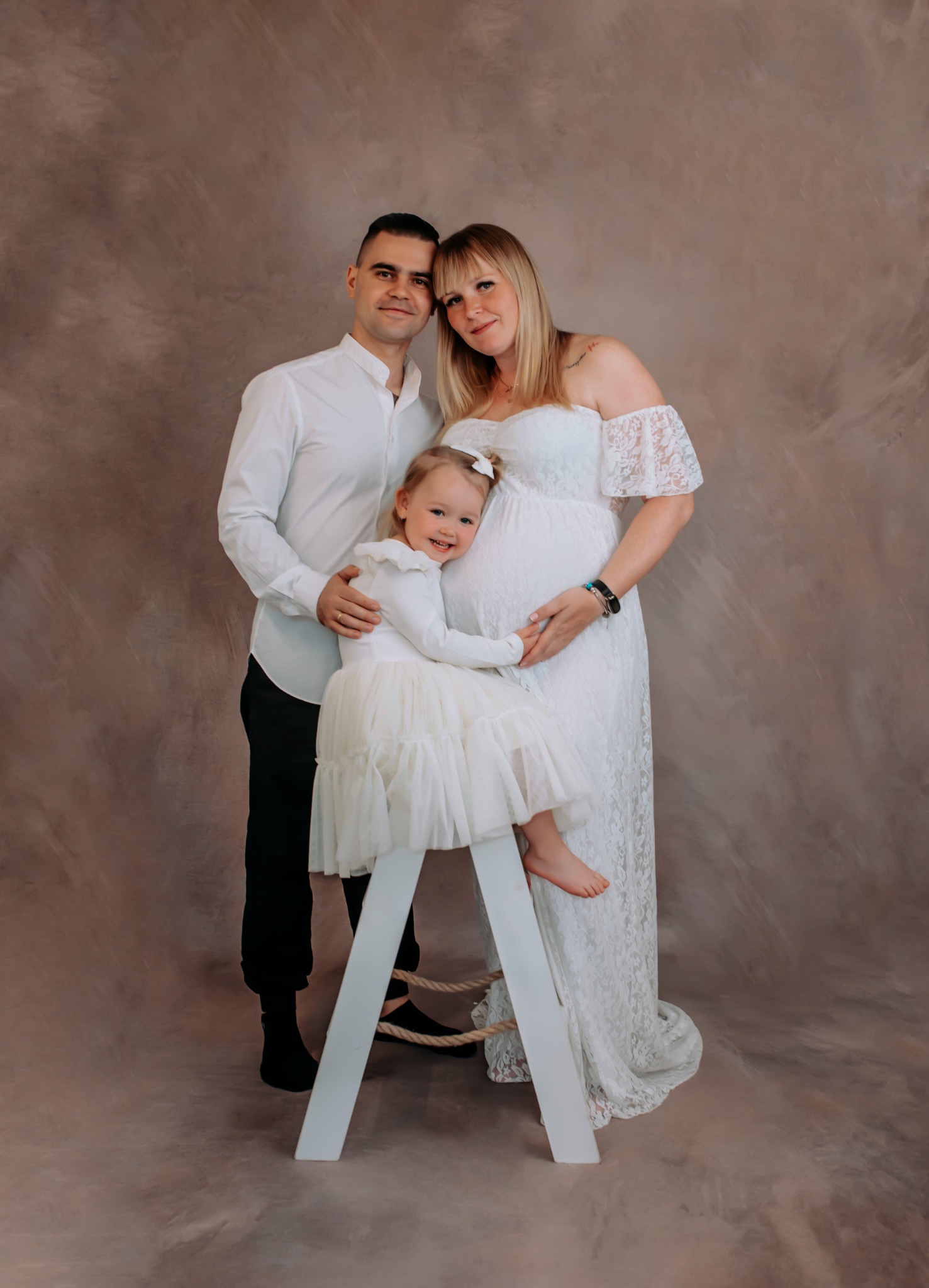 Uśmiechnięta rodzina: tata obejmuje mamę w ciąży, a córka siedzi na białej drabince, wszyscy ubrani na biało na tle abstrakcyjnego tła.