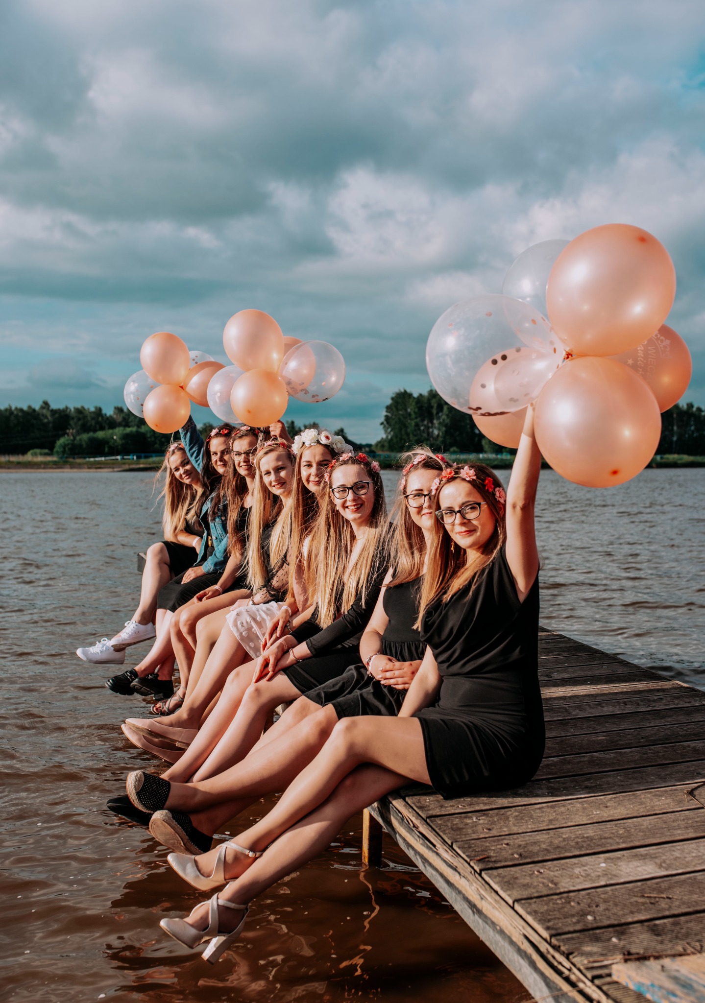 Grupa młodych kobiet siedzi na drewnianym pomoście nad wodą, trzymając różowe i przezroczyste balony, ubrane w sukienki i wianki z kwiatów.