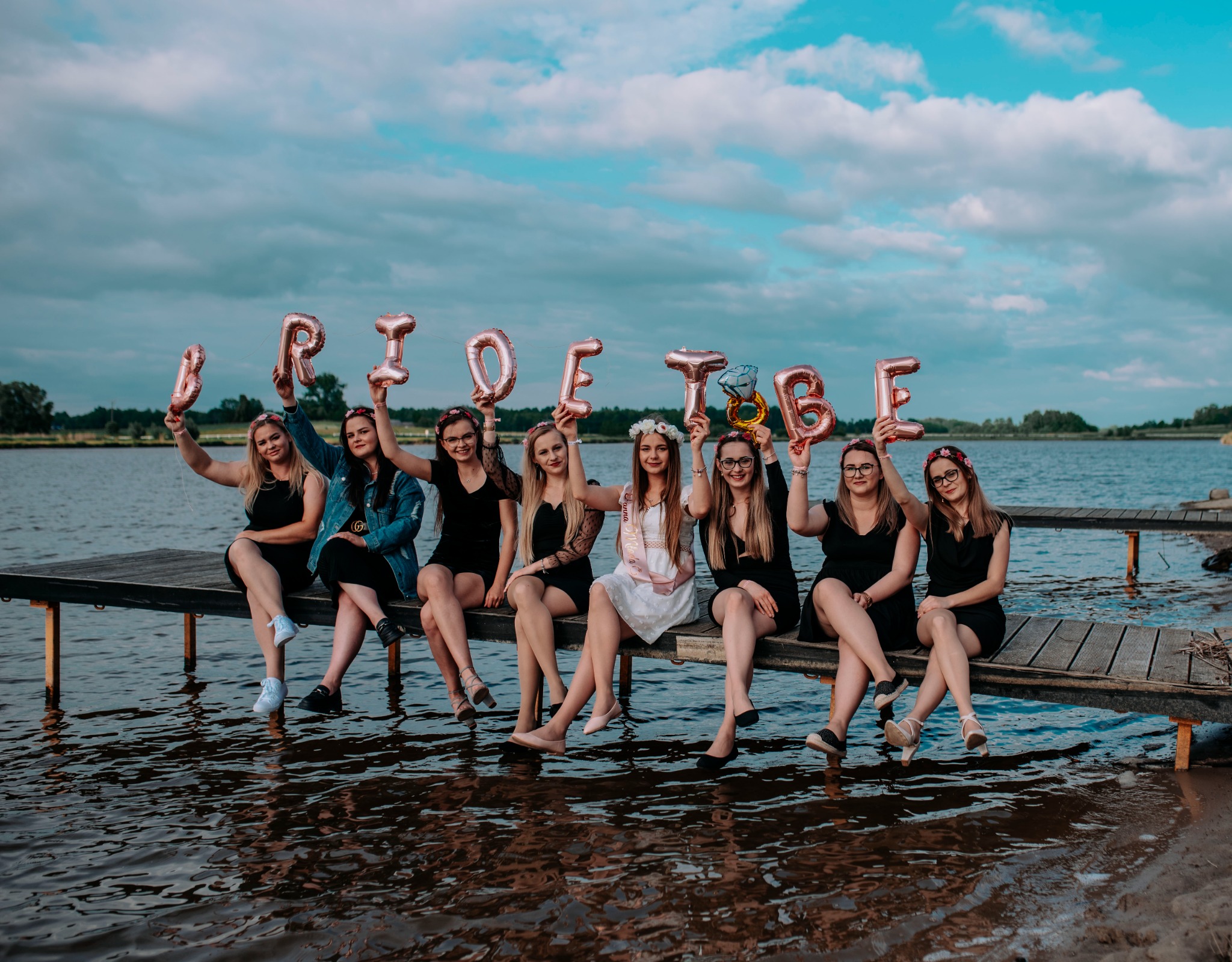 Grupa kobiet siedzi na drewnianym pomoście nad jeziorem, trzymając balony w kształcie napisu 'BRIDETOBE'; panna młoda w białej sukience z szarfą.